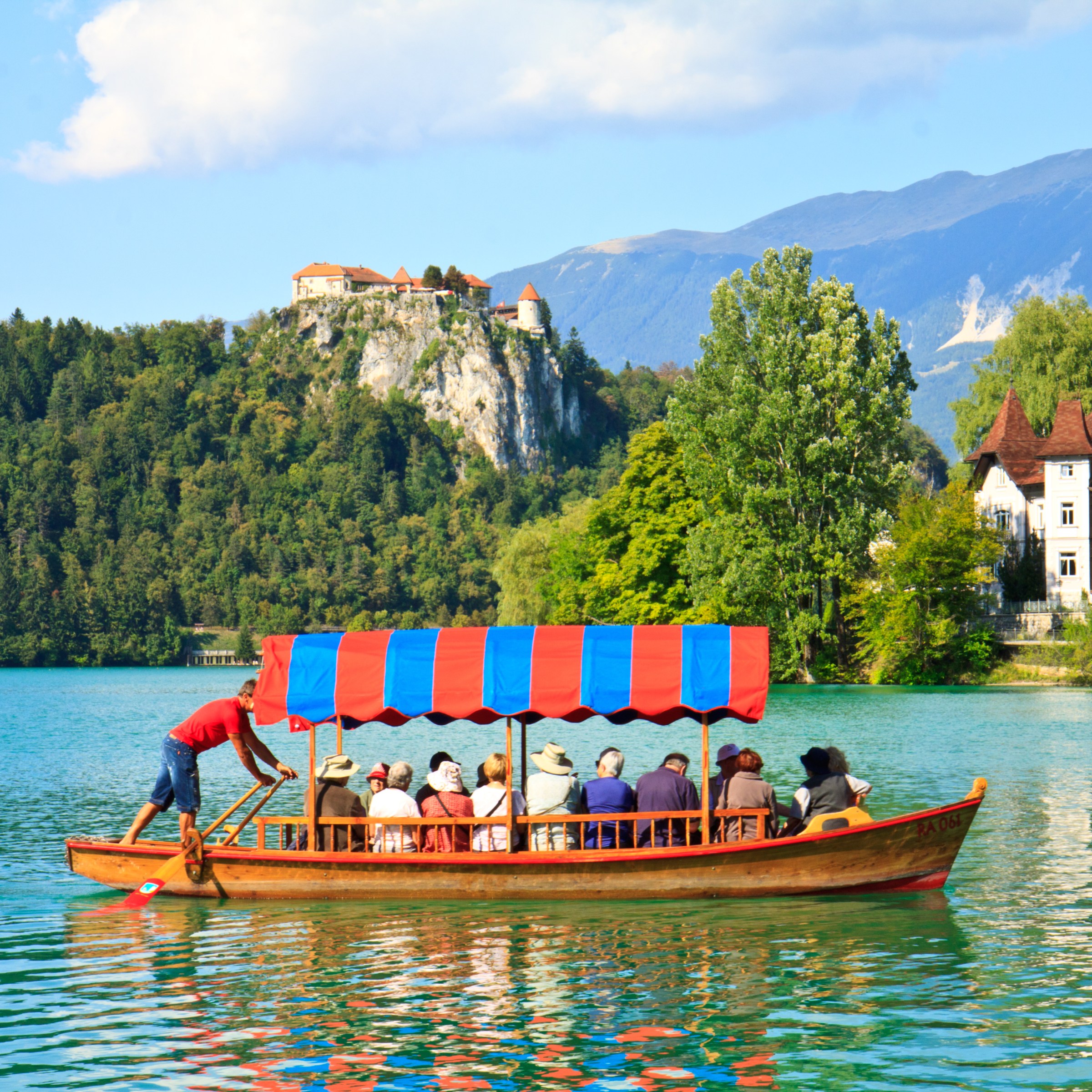 People in a wooden boat with a red-blue canopy on a lake, castle and mountains in the background.