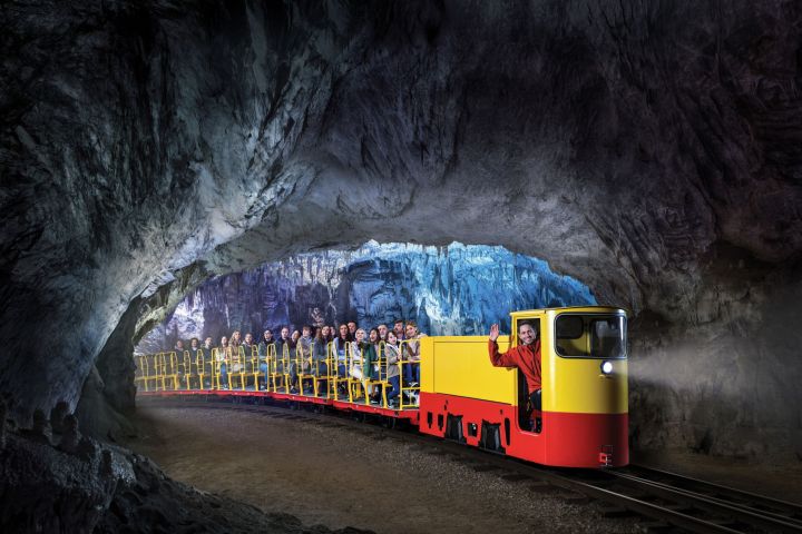 Tourist train with passengers inside a large, illuminated cave tunnel.