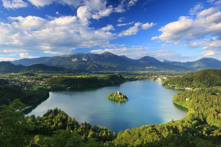 Lake with island church, surrounded by lush green hills and distant mountains under a partly cloudy sky.
