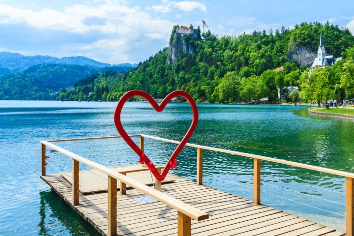 Red heart sculpture on a wooden pier by a calm lake with a castle on a hill in the background.