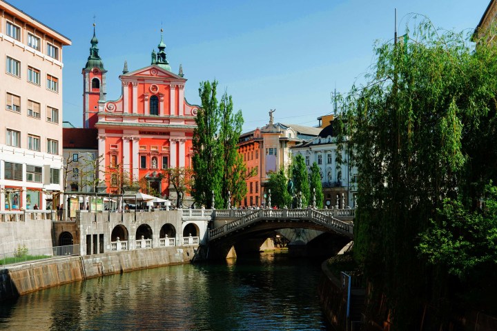 River view with a pink church, arched bridge, and trees in a European city landscape.