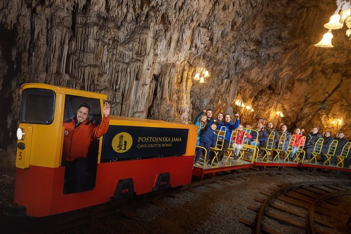 People on a yellow train inside Postojna Cave with stalactites and lit chandeliers above.
