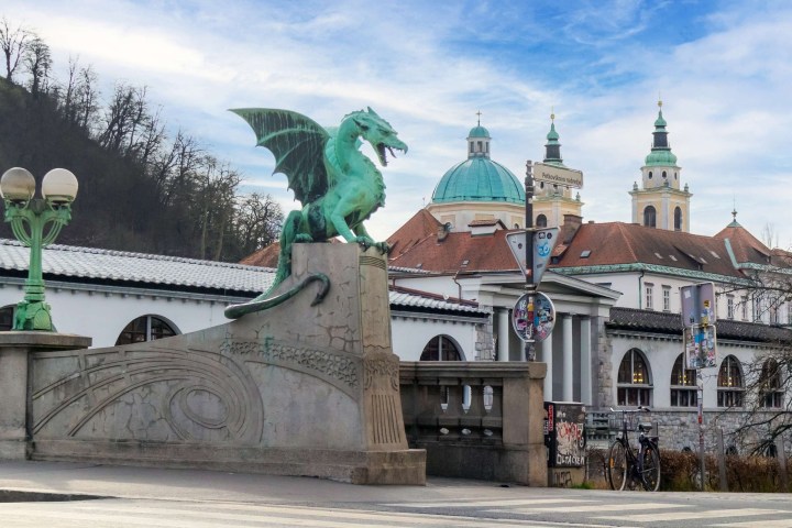 Green dragon statue on a bridge with buildings and domes in the background under a blue sky.