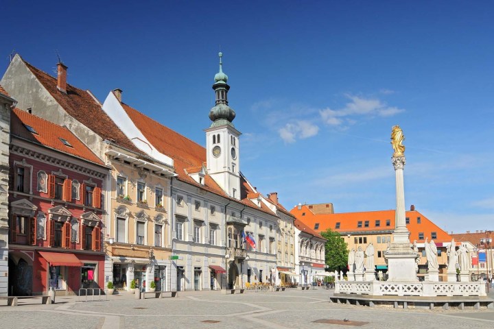 Historic European square with colorful buildings and ornate column sculpture under a blue sky.