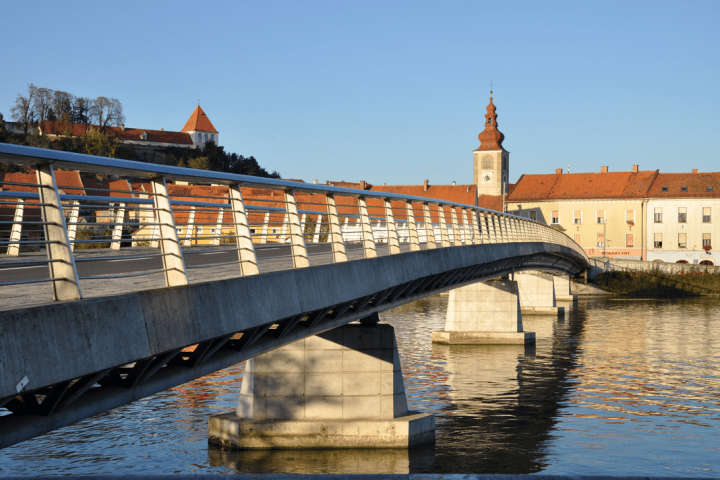 Pedestrian bridge over a river, with a clock tower and buildings in the background.