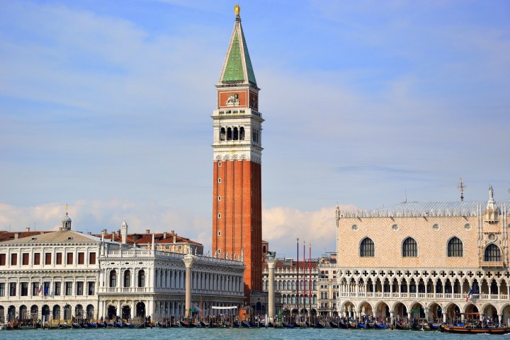 Tall red bell tower and historic buildings by a canal under a blue sky.