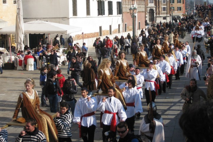 People in period costumes parade through a crowd-lined street in a historic European city.