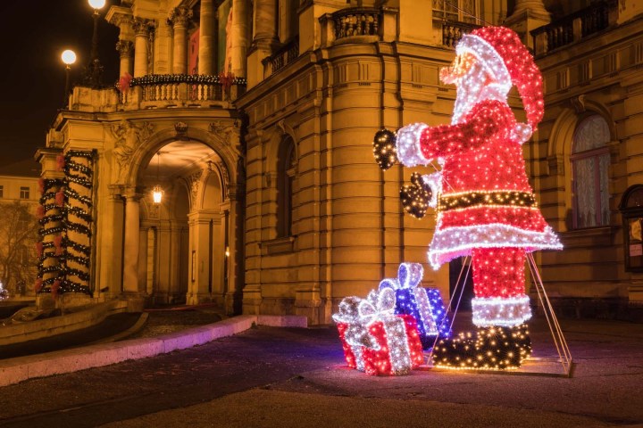 Santa Claus lights display with gift boxes in front of a lit historical building at night.