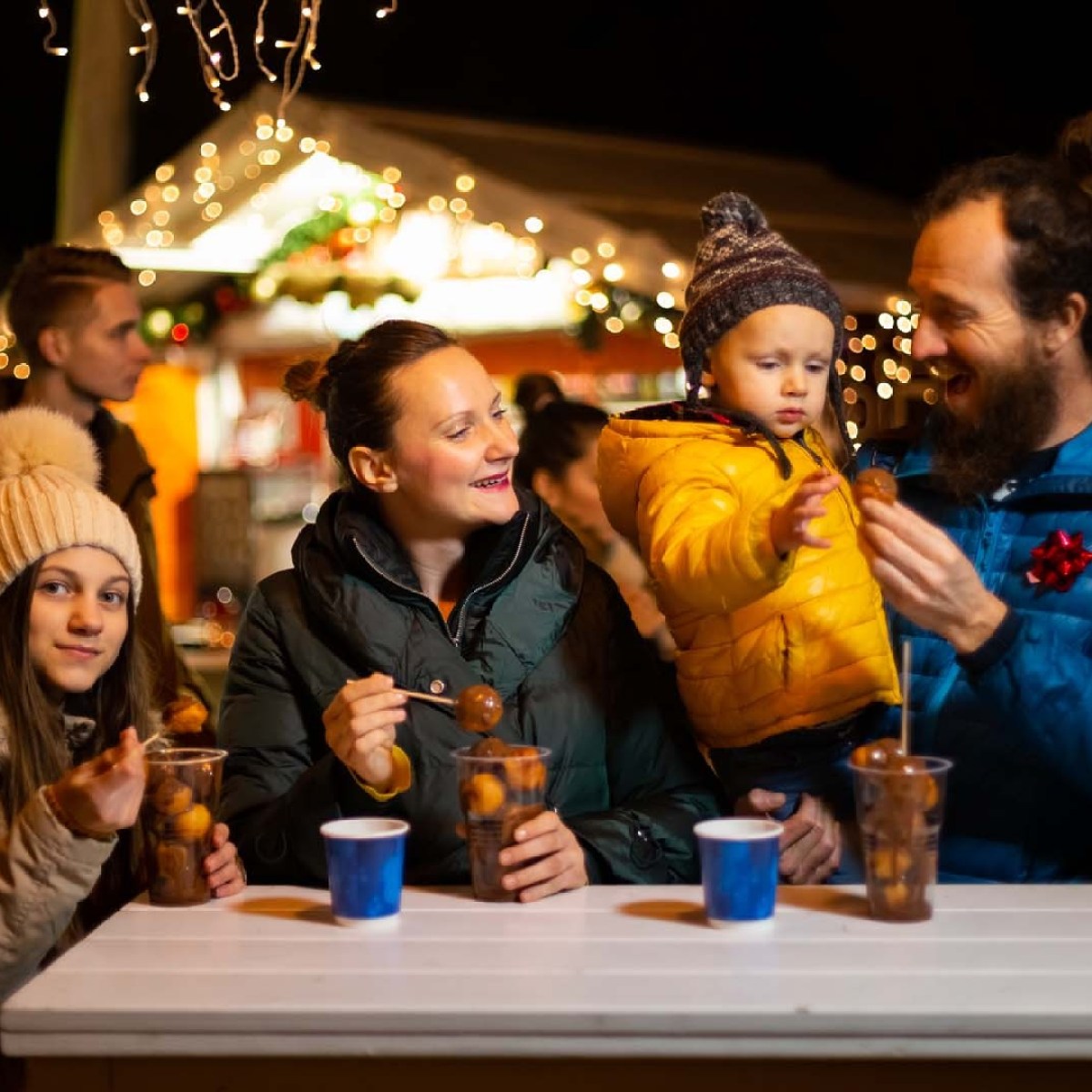 Family enjoying warm drinks and snacks at a festive outdoor market with lights.