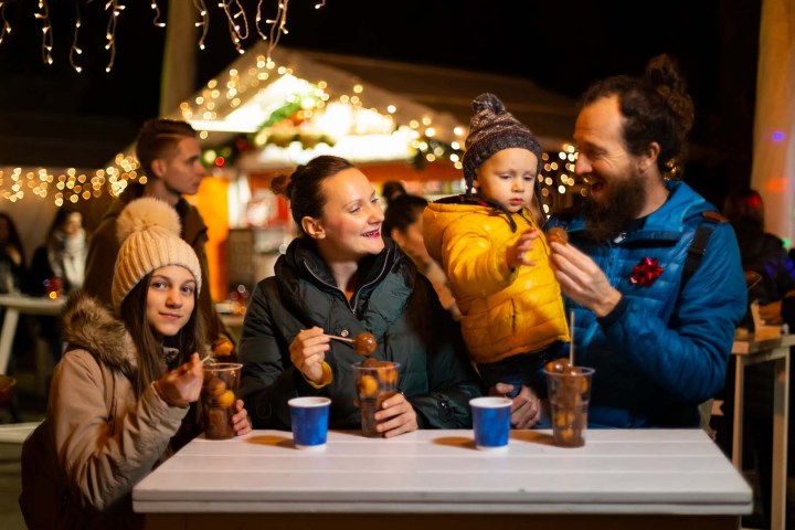 Family enjoying warm drinks and snacks at a festive outdoor market with lights.
