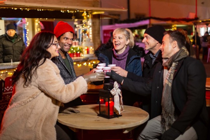 Group of people at a festive market, toasting with drinks around a wooden table.