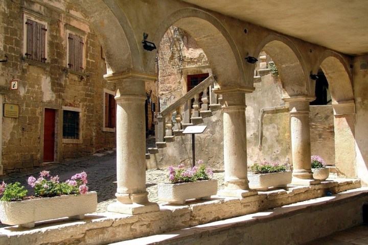 Stone archways with plant boxes and flowers overlooking a rustic courtyard with stairs and old stone buildings.