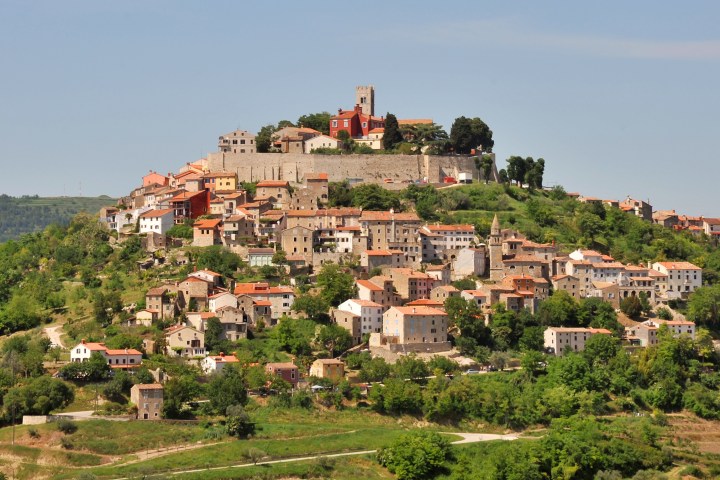 Hilltop town with stone houses and a tower, surrounded by greenery under a clear blue sky.