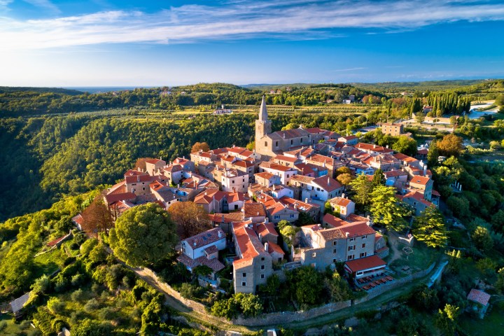 Aerial view of a quaint village with red-roofed buildings and a central church, surrounded by lush green landscape.