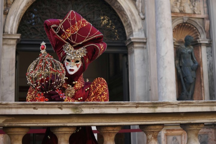 Person in colorful Venetian carnival costume and mask holding a decorative orb on a stone balustrade.
