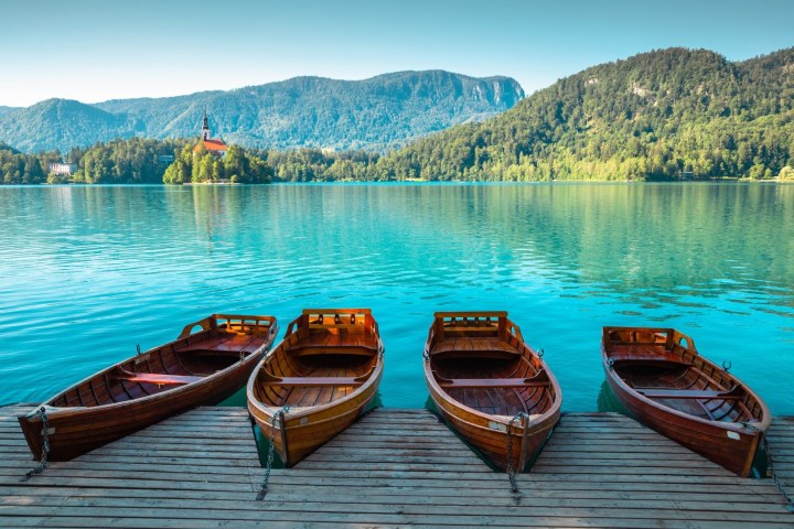 Four empty wooden boats docked on clear lake with mountainous forest backdrop.