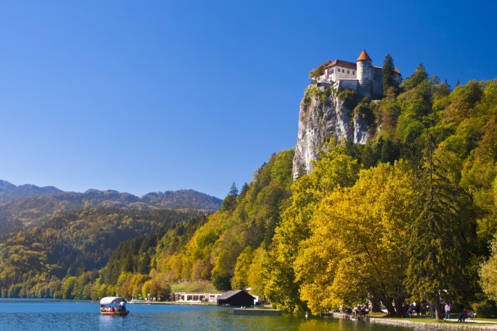 Castle on a cliff above a lake with autumn trees and a boat in the water.