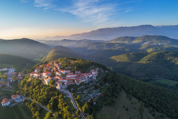 Aerial view of a hilltop town with red roofs surrounded by green landscape and distant mountains under a clear sky.