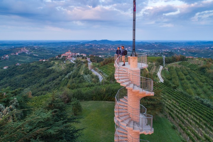 Two people stand on a spiral tower overlooking a hilly vineyard landscape at dusk.
