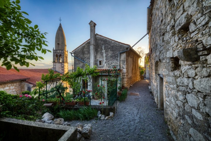 Stone pathway by old buildings and bell tower at sunset, lush greenery.