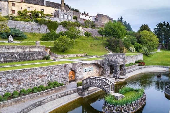 Historic castle with stone bridge over moat surrounded by greenery and gardens.