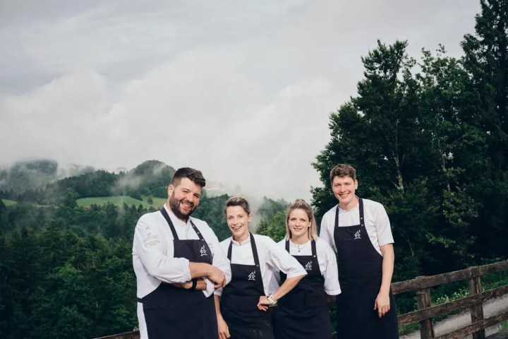 Four chefs in black aprons smiling outdoors with a forest background.