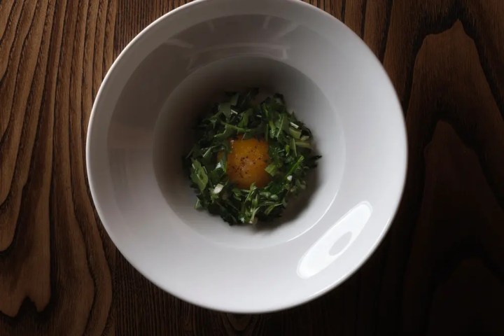 White bowl with greens and egg yolk on a wooden table.