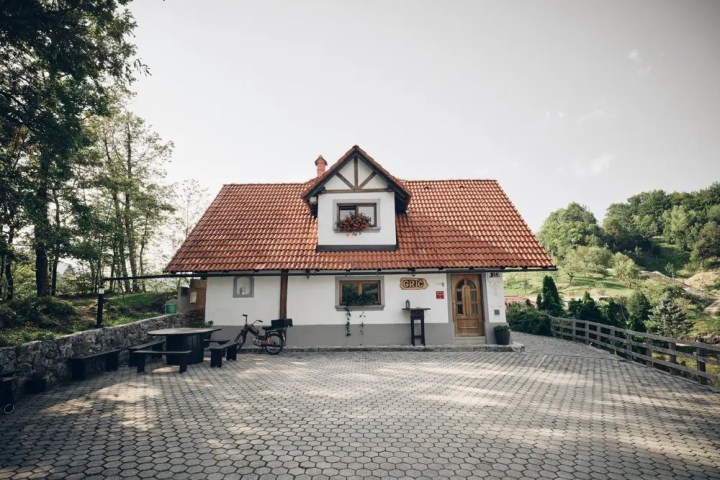 A small, white house with a red roof and a cobblestone patio surrounded by greenery.