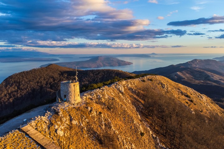 Hilltop stone tower overlooking scenic landscape with sea and mountains at sunset.