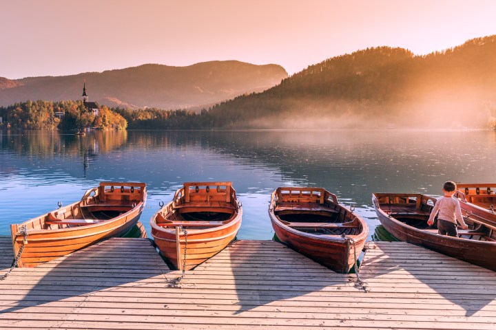 Five wooden boats docked on a serene lake with misty mountains in the background at sunrise.