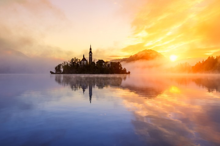 Serene lake at sunrise with island and church silhouette, surrounded by mist.