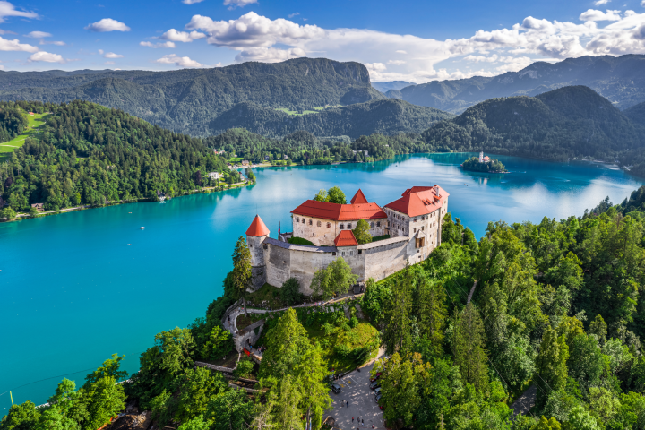 Aerial view of a castle on a hill by a vibrant blue lake surrounded by mountains and trees under a partly cloudy sky.