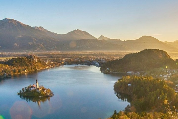 A serene lake with an island church, surrounded by mountains under a clear sky at sunset.
