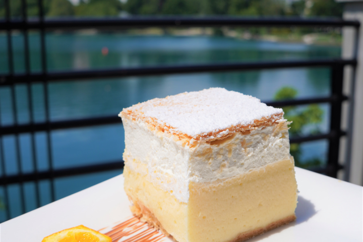 Square dessert with cream layers on plate, orange slice garnish, scenic lake view background.