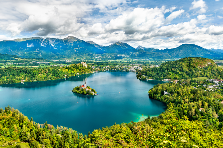 Scenic view of a lake with an island, surrounded by mountains and greenery under a cloudy sky.