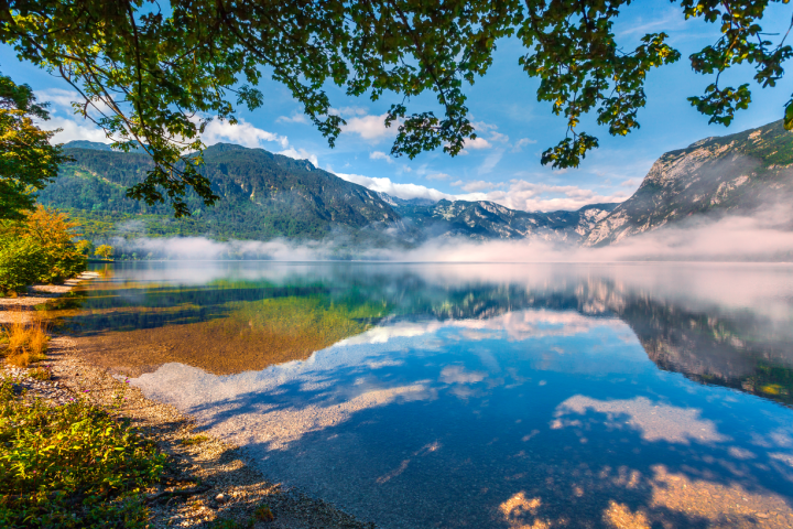Serene lake with mist, trees, and mountains under a clear blue sky.