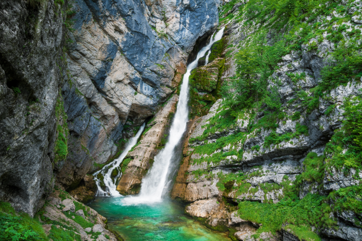 Waterfall cascading down a rugged cliff into a turquoise pool, surrounded by lush green foliage.