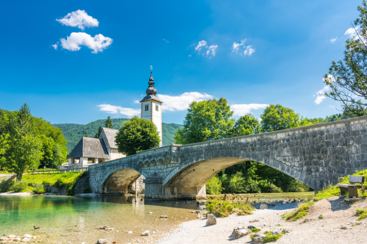 Stone bridge and church tower over a river, surrounded by trees and mountains under a clear blue sky.
