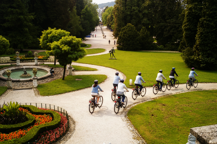 Cyclists on a park path surrounded by greenery and a circular fountain.