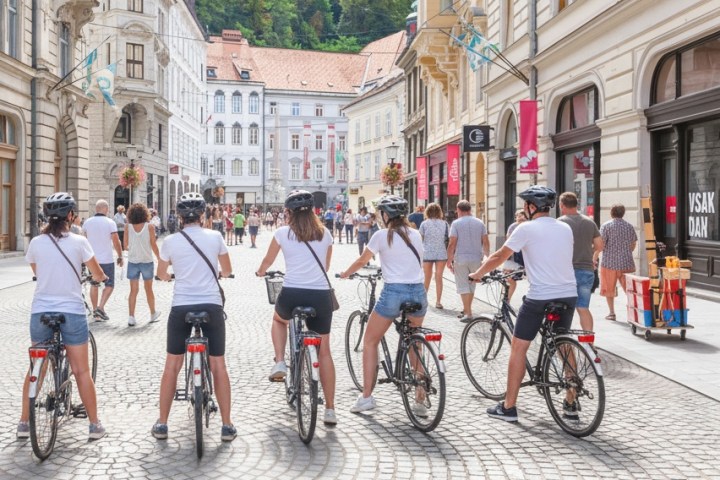 Group of cyclists wearing helmets in a European city street, surrounded by pedestrians and historic buildings.