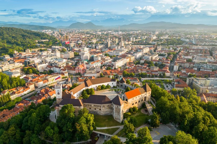 Aerial view of a hilltop castle surrounded by a cityscape with distant mountains under a cloudy sky.