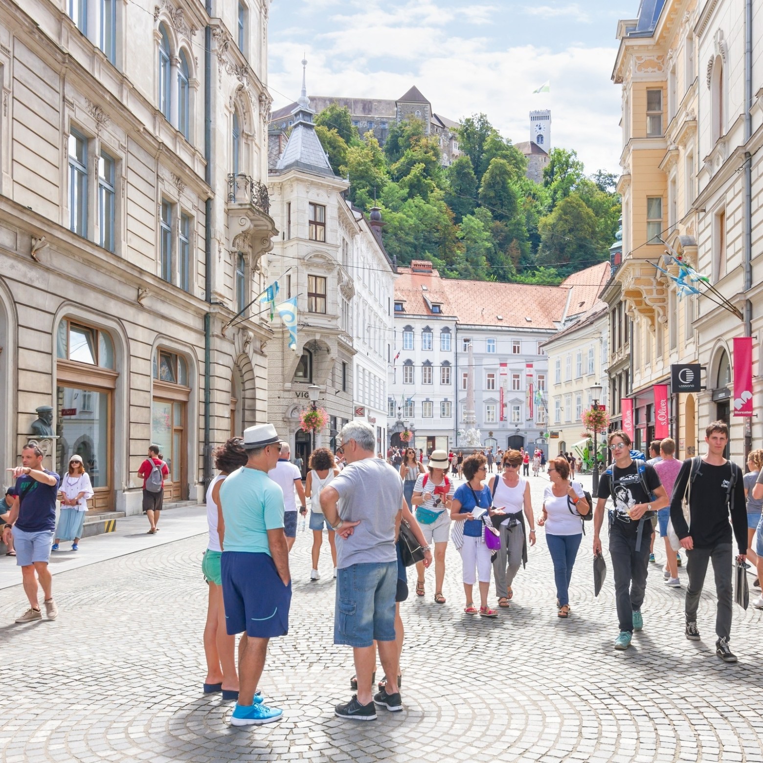 Crowded street with historic buildings and a hilltop castle in the background, people walking and shopping.