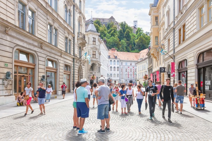 Crowded street with historic buildings and a hilltop castle in the background, people walking and shopping.