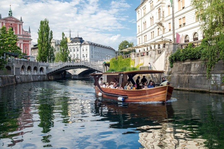 Wooden boat with passengers on a canal with historic buildings and a bridge in the background.