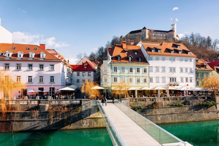 Colorful riverside buildings, bridge, and castle on hill under clear sky.
