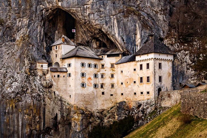 Castle built into a cliffside with cave entrance above, surrounded by rocky terrain.