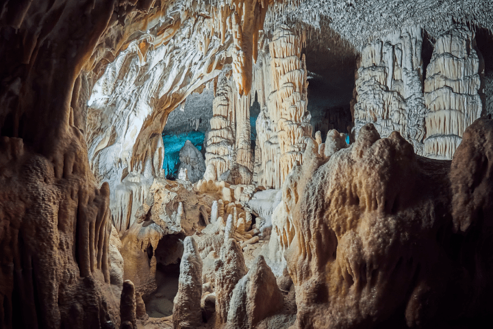 A cave interior with stalactites and stalagmites in earthy tones, illuminated by blue and white light.