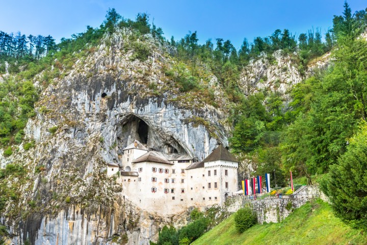A castle built into a rocky hillside with large cave, surrounded by trees.