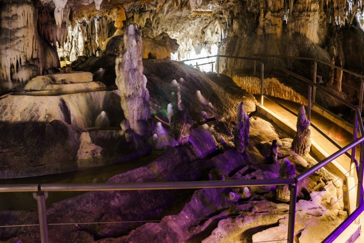 Lit cave with stalactites, stalagmites, and a walkway with railings.