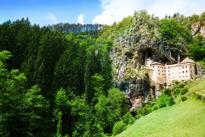 A cliffside castle partially built into a rocky mountain, surrounded by dense green forest.